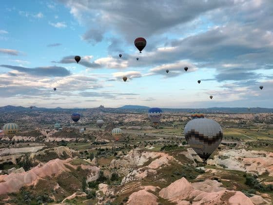 Numerosos globos aerostáticos flotando sobre un vasto paisaje de valle rocoso bajo un cielo parcialmente nublado.