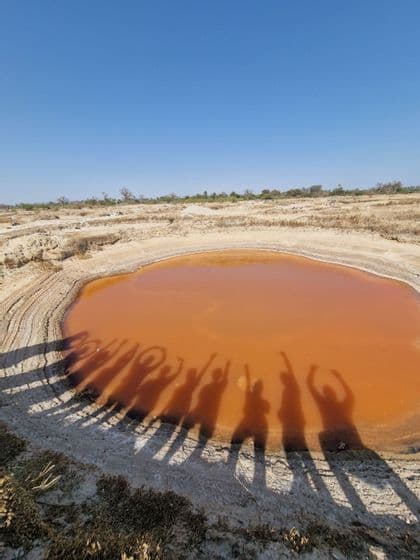 Ombre di un gruppo WeRoad in viaggio che posano con le braccia alzate sopra una piccola piscina arancione in un paesaggio arido.