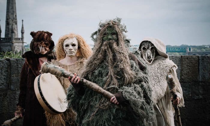 Quattro persone in elaborati costumi e maschere folkloristiche, con un tamburo e bastoni, posano all'aperto vicino a un muro di pietra.