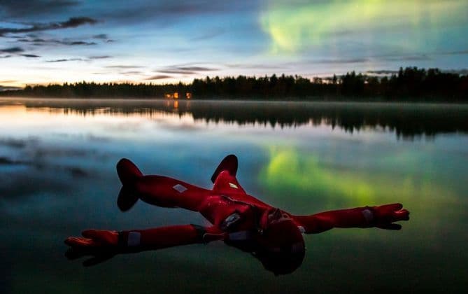 Una persona con un traje de supervivencia rojo flota de espaldas en un lago en calma, con la aurora boreal reflejada en el agua al atardecer.