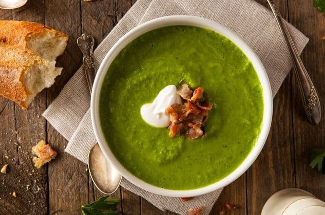 Un bol de soupe verte garni de crème et de lardons est posé sur une table en bois, à côté d'un morceau de pain croustillant et d'une cuillère.
