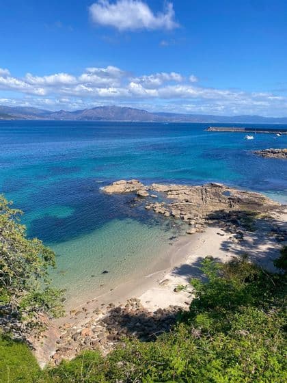Veduta dall'alto di una costa rocciosa e una piccola spiaggia sabbiosa, con acqua turchese cristallina e montagne lontane oltre il mare.