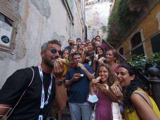 A WeRoad group trip smiles for the camera while eating cannoli on a narrow outdoor staircase.