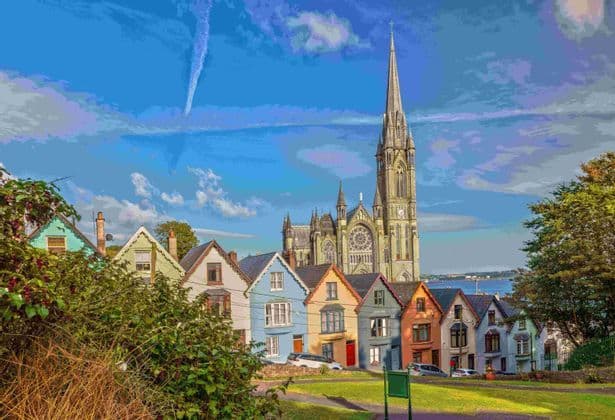 Une rangée de maisons colorées se trouve devant une grande cathédrale gothique en pierre, sous un ciel bleu parsemé de nuages.
