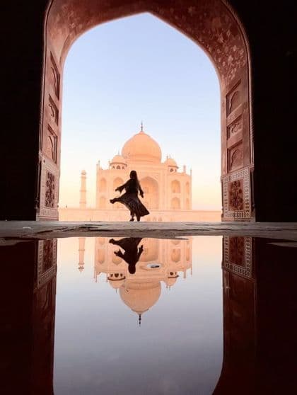 Una persona in silhouette posa davanti al Taj Mahal, ripresa attraverso un arco e con il suo riflesso in una pozzanghera in primo piano.
