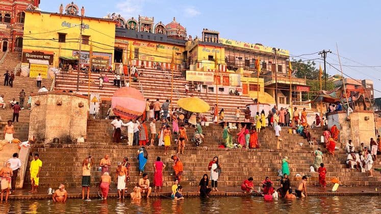 Una grande folla di persone in abiti colorati si raduna sui ghat di pietra, con molti che si bagnano nel fiume sotto tradizionali edifici gialli.