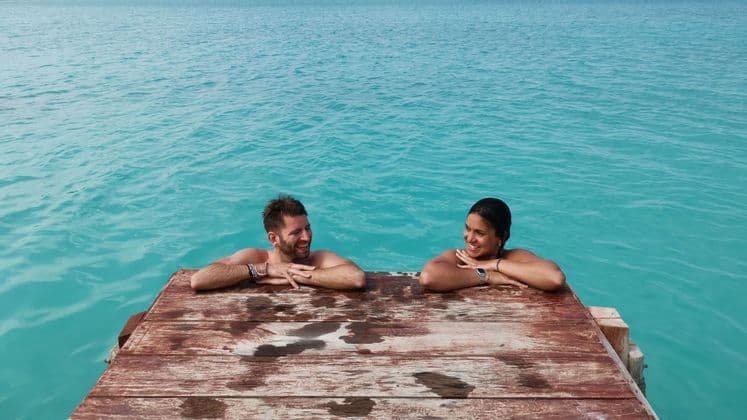 Ein Mann und eine Frau auf einer WeRoad-Gruppenreise lehnen an einem Holzsteg und lächeln sich im türkisfarbenen Wasser an.
