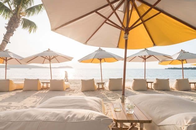 Sous un grand parasol jaune et blanc, des poufs sont installés sur une plage de sable face à la mer au lever du soleil.