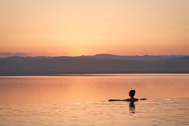 Una persona vista di schiena si rilassa in una piscina a sfioro che si affaccia su un grande specchio d'acqua e montagne distanti durante un caldo tramonto.