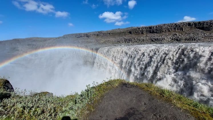 Un arcobaleno si inarca sulla nebbia di una potente cascata che precipita in un canyon roccioso sotto un cielo azzurro e limpido.