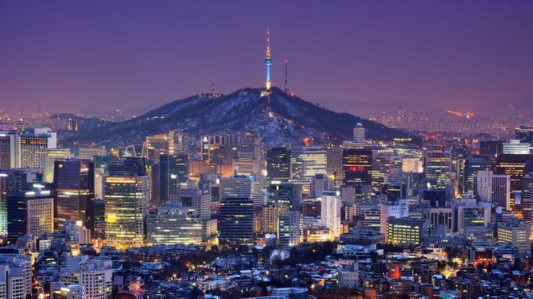 A sprawling city skyline illuminated at twilight, with a tall tower on a snow-dusted mountain in the background.