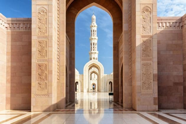 Un minaret de mosquée est encadré par une grande arche en pierre surplombant une cour en marbre poli, sous un ciel bleu clair.