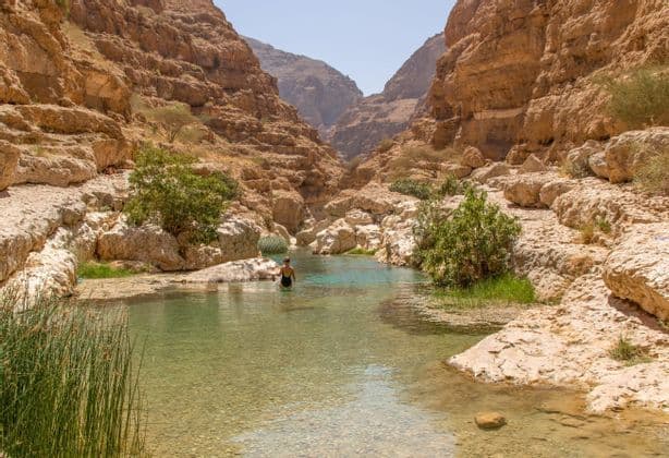 Une personne en maillot de bain noir patauge dans un ruisseau clair au cœur d'un canyon rocheux et ensoleillé.