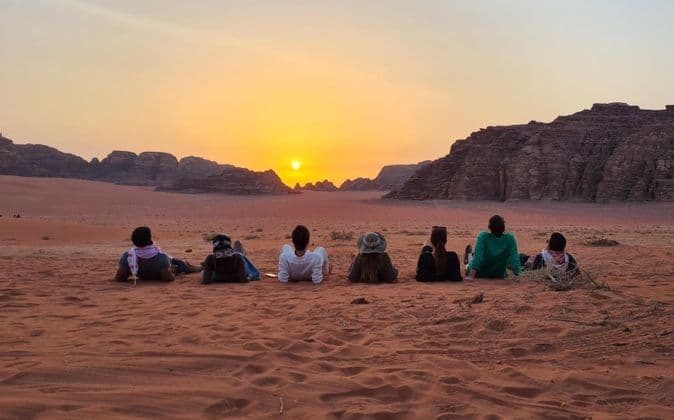 Un grupo de WeRoad en un viaje contempla el atardecer sobre la arena roja del desierto, con formaciones rocosas distantes al fondo.