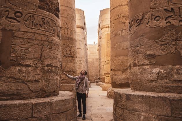 Ein Mann steht in einer alten Tempelhalle und blickt zu den Hieroglyphen auf, die in die massiven Steinsäulen gemeißelt sind.