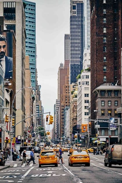 A busy city street with yellow taxis driving and pedestrians crossing, flanked by tall skyscrapers under a clear sky.
