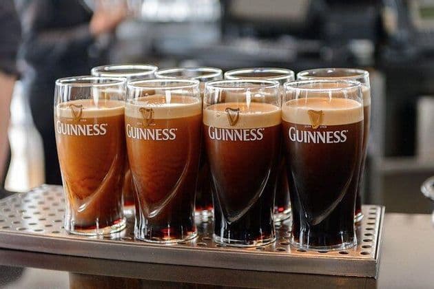 Due file di bicchieri da pinta Guinness pieni di stout scura e schiuma cremosa si trovano su un vassoio di metallo.