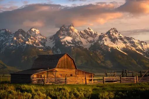 Un fienile in legno con una recinzione si trova in un campo erboso di fronte a una catena montuosa innevata sotto un cielo nuvoloso e illuminato dal sole.