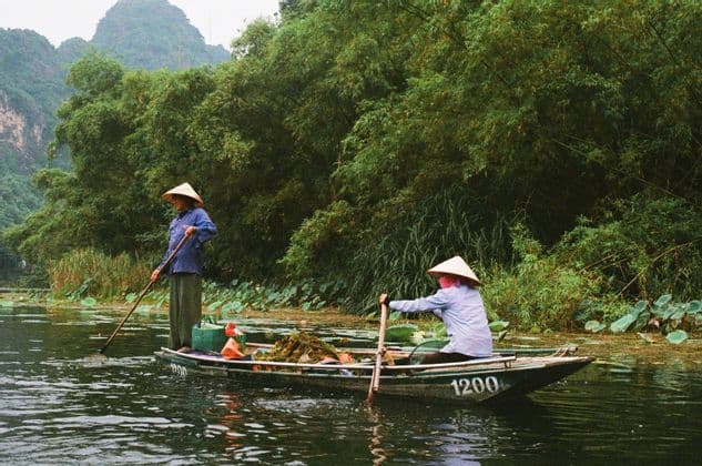 Deux personnes aux chapeaux coniques naviguent sur une rivière entourée d'une végétation luxuriante et de collines, l'une à la perche et l'autre à la pagaie.