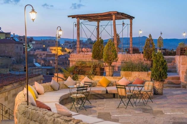 Une terrasse de toit en pierre avec des banquettes capitonnées, des tables et des lampadaires éclairés, offrant une vue sur un village à flanc de colline au crépuscule.