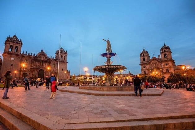 Una plaza histórica al atardecer con una fuente central ornamentada, flanqueada por dos catedrales iluminadas mientras la gente camina.