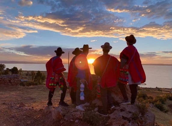 Eine WeRoad-Gruppe mit roten Ponchos und Hüten beobachtet den Sonnenuntergang von einem Hügel mit Blick auf einen großen See.