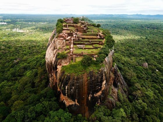 Aerial view of a large rock fortress with ancient ruins on its summit, surrounded by a vast, dense green jungle.