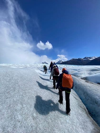 A WeRoad group trip with backpacks and crampons hiking in a line across a vast, sunlit glacier under a blue sky.