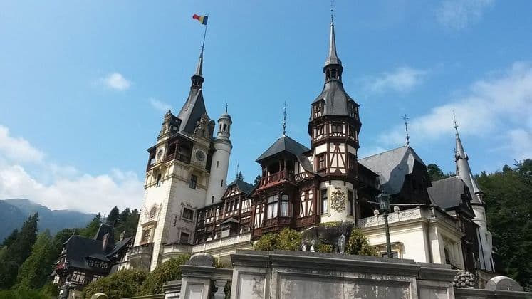 Un château orné avec plusieurs tours et flèches, situé devant une montagne boisée, avec un drapeau flottant au sommet de la plus haute flèche.
