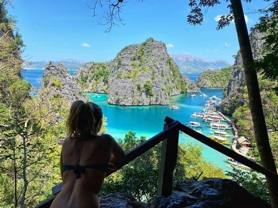 Eine Frau im Bikinioberteil blickt von einem erhöhten Standpunkt über eine türkisfarbene Lagune mit vielen Booten und felsigen, grünen Inseln.