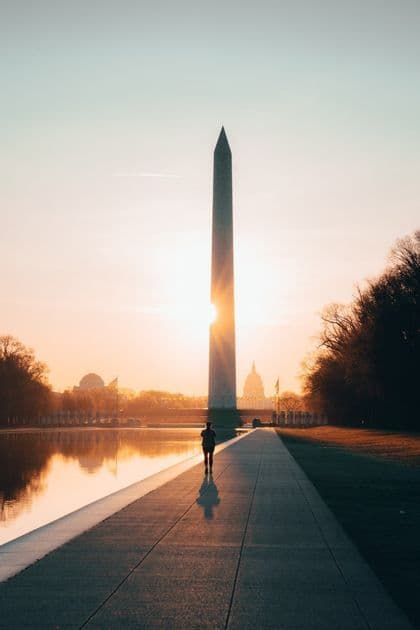 A person walks on a path beside a reflecting pool towards a tall obelisk as the sun flares from behind it at sunrise.
