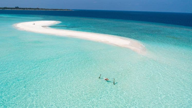 Vista aerea di una persona che galleggia su una zattera in acque cristalline turchesi, accanto a un sinuoso banco di sabbia bianco e all'oceano blu profondo.