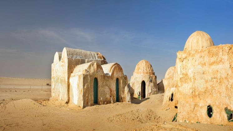 Diversi edifici in stile adobe con tetti a cupola si ergono in un deserto sabbioso sotto un cielo azzurro e limpido.