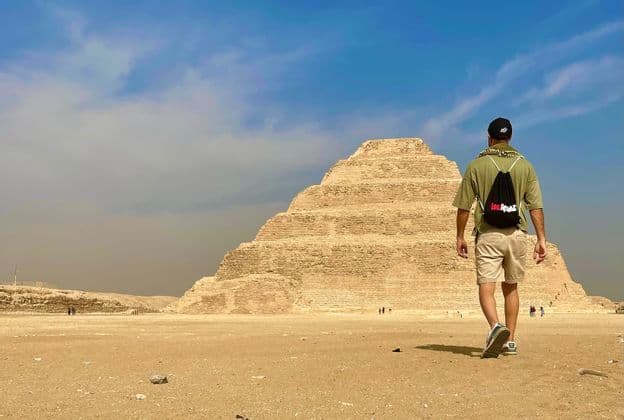 Un viaggiatore con uno zaino WeRoad attraversa un paesaggio desertico sabbioso verso una grande piramide a gradoni.