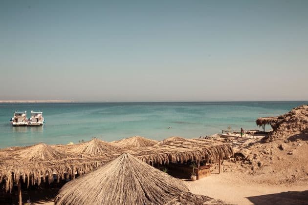 Thatched-roof umbrellas on a sandy shore with two boats anchored in calm, turquoise water under a clear sky.