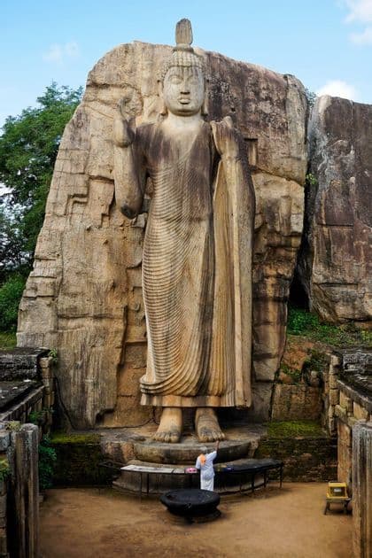 A person in white clothing stands at the base of a colossal Buddha statue carved from a rock cliff, viewed from above.