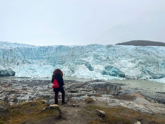 Una persona con uno zaino rosso si trova su una riva rocciosa, guardando l'immensa parete di un ghiacciaio sotto un cielo grigio.