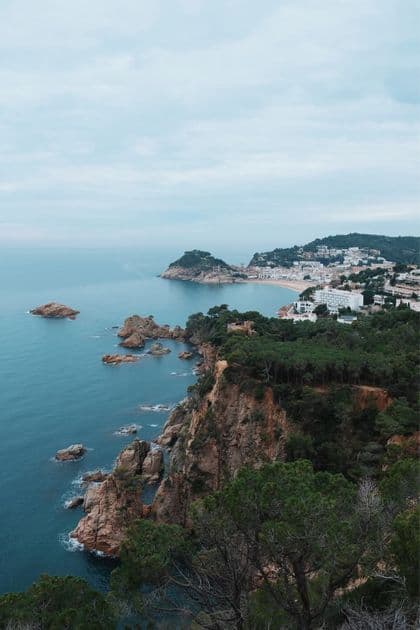 Vue plongeante d'une côte rocheuse et arborée menant à une ville balnéaire avec une plage de sable sous un ciel nuageux.