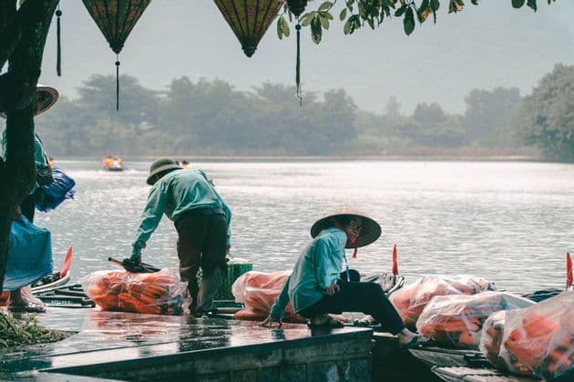 Due persone con cappelli conici che sistemano giubbotti di salvataggio arancioni su un pontile di legno bagnato vicino a un fiume, con lanterne appese a un albero.