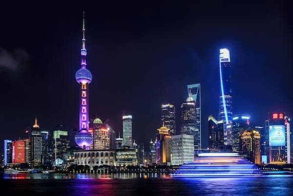 Uno skyline cittadino con un'alta torre sferica illuminata di viola di notte, con una barca che lascia scie luminose sull'acqua di fronte.