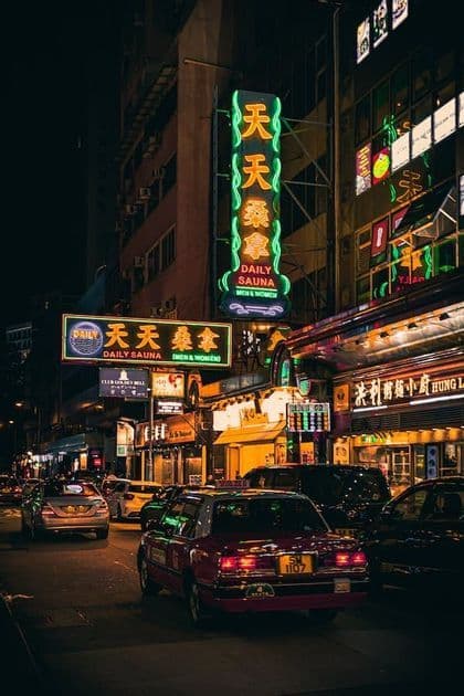 Una via cittadina animata di notte, affollata di auto e illuminata da grandi insegne al neon luminose con caratteri asiatici.