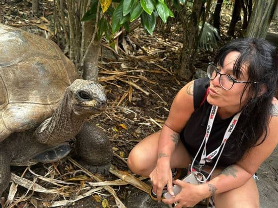 Una donna con occhiali e un laccetto WeRoad si accovaccia faccia a faccia con una tartaruga gigante in una foresta.