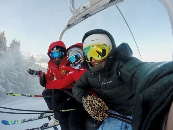 Drei Personen einer WeRoad-Gruppe in Skiausrüstung machen ein Selfie auf einem Skilift vor verschneiter Bergkulisse.