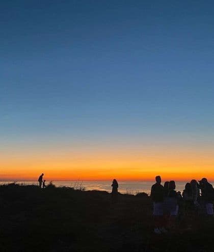 Sagome di un viaggio di gruppo WeRoad su una collina che osservano il tramonto arancione e blu sul mare.