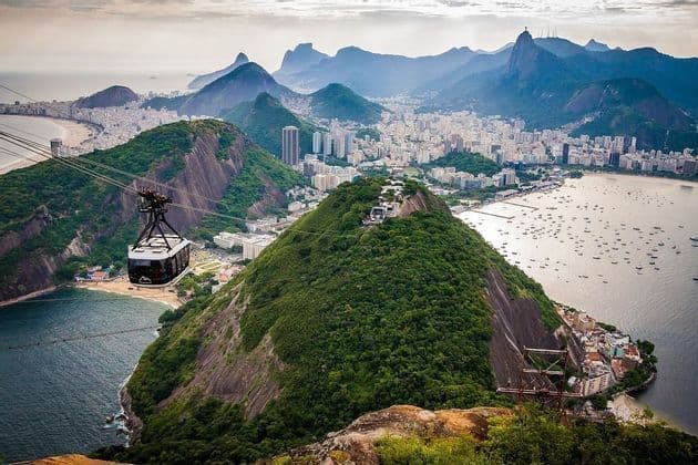 Un teleférico viaja sobre una exuberante montaña verde, con una vista panorámica de una ciudad costera, una bahía con barcos y montañas distantes.