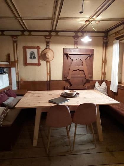 Interno di una stanza rustica con un tavolo di legno, due sedie e una panca, contro pareti rivestite in legno e un soffitto a cassettoni.