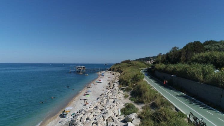 Panoramica aerea di una costa rocciosa, con spiaggia, pista ciclabile verde e un trabucco in legno che si protende nel blu del mare.