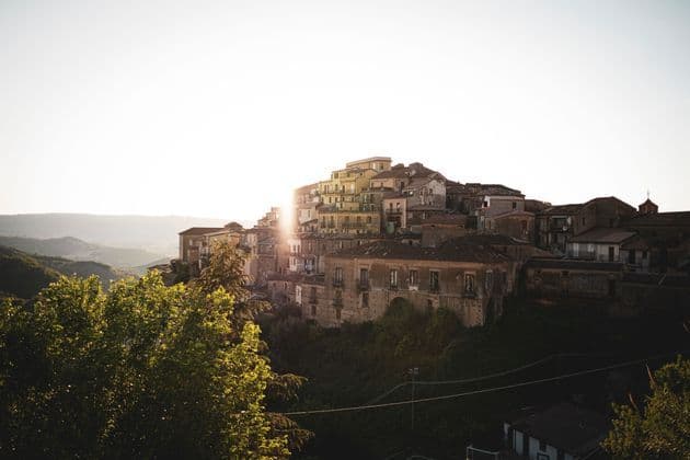 Un borgo di vecchie case costruite su un fianco di collina verdeggiante, con il sole nascente che crea un bagliore luminoso da dietro gli edifici.