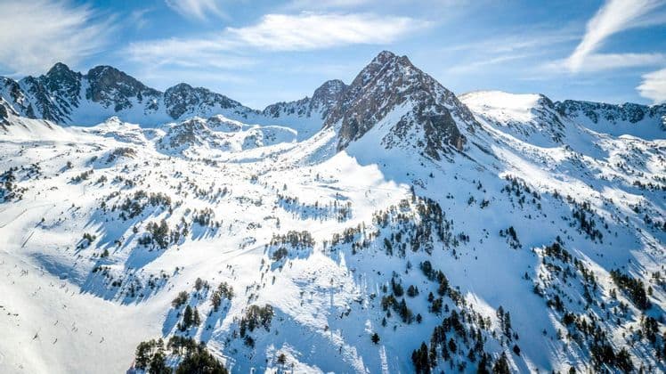 Una vasta cordillera cubierta de nieve, salpicada de árboles perennes bajo un cielo azul brillante con nubes tenues.