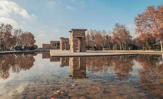 Un tempio di pietra con grandi portali si riflette in uno specchio d'acqua tranquillo, circondato da alberi con fogliame autunnale.